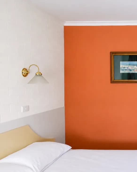 A queen bed against an orange feature wall, with a brass wall lamp and a framed print.