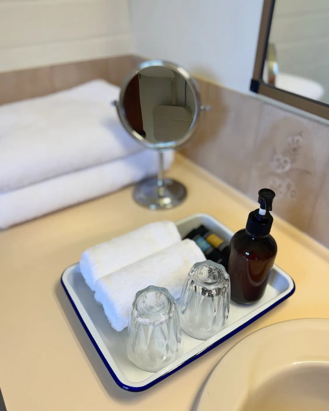 Bathroom tray with rolled white towels, two glasses and a bottle of hand wash beside a round mirror.