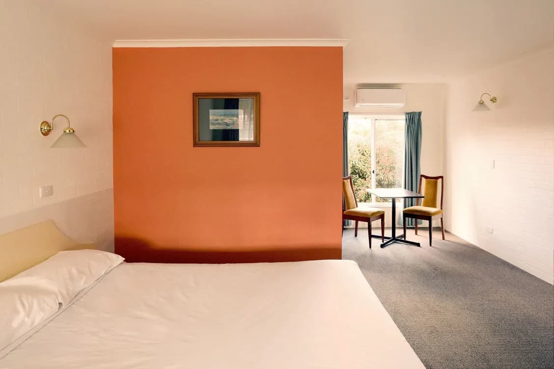 Bedroom with bed, orange accent wall, table and chairs by a window.