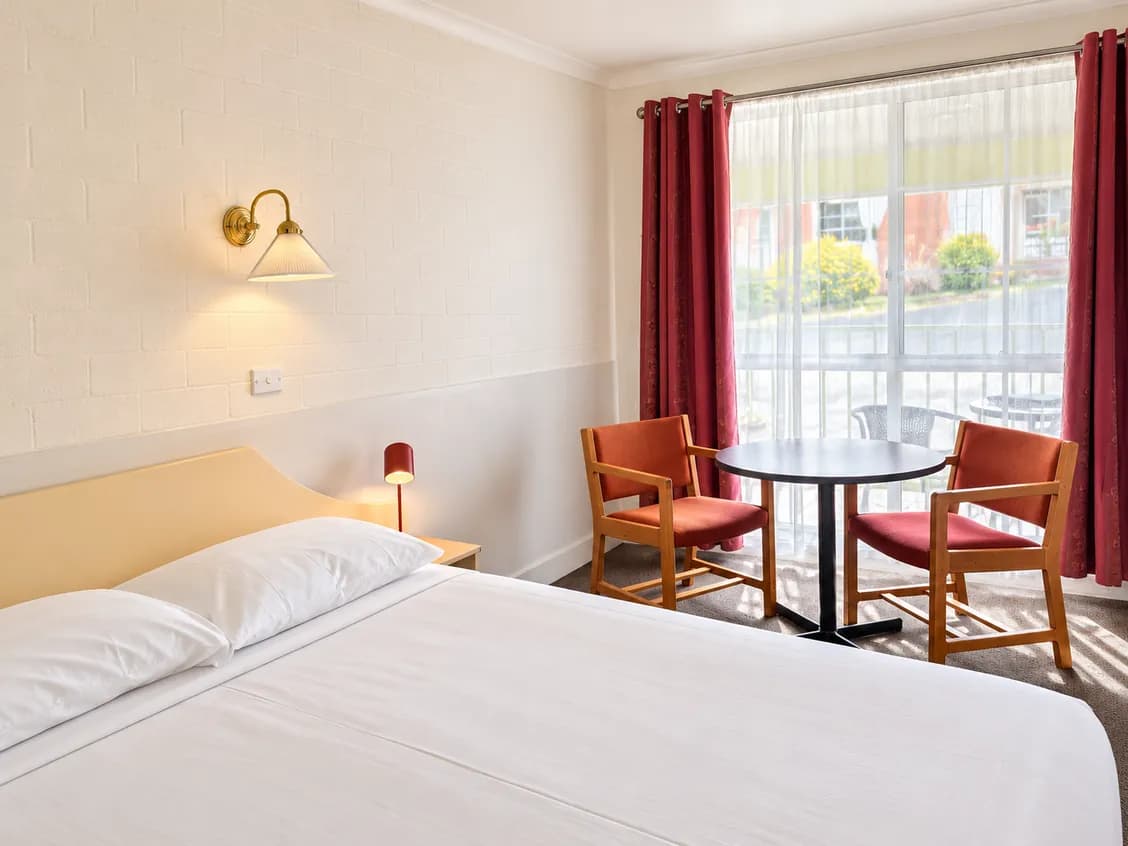 Bed with white linen, a brass wall lamp, and a round table and two red chairs by a window with red curtains.