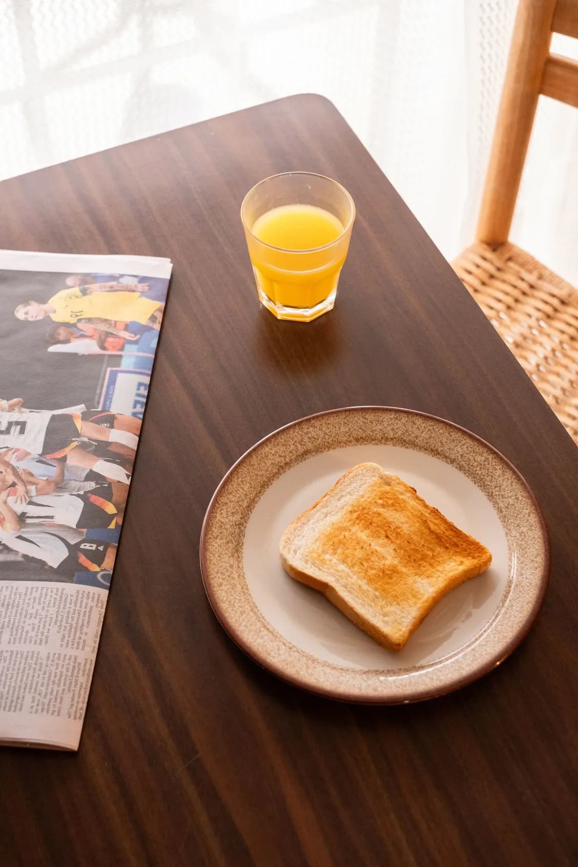 Toast on a plate, glass of orange juice, and newspaper on a wood table near a window.