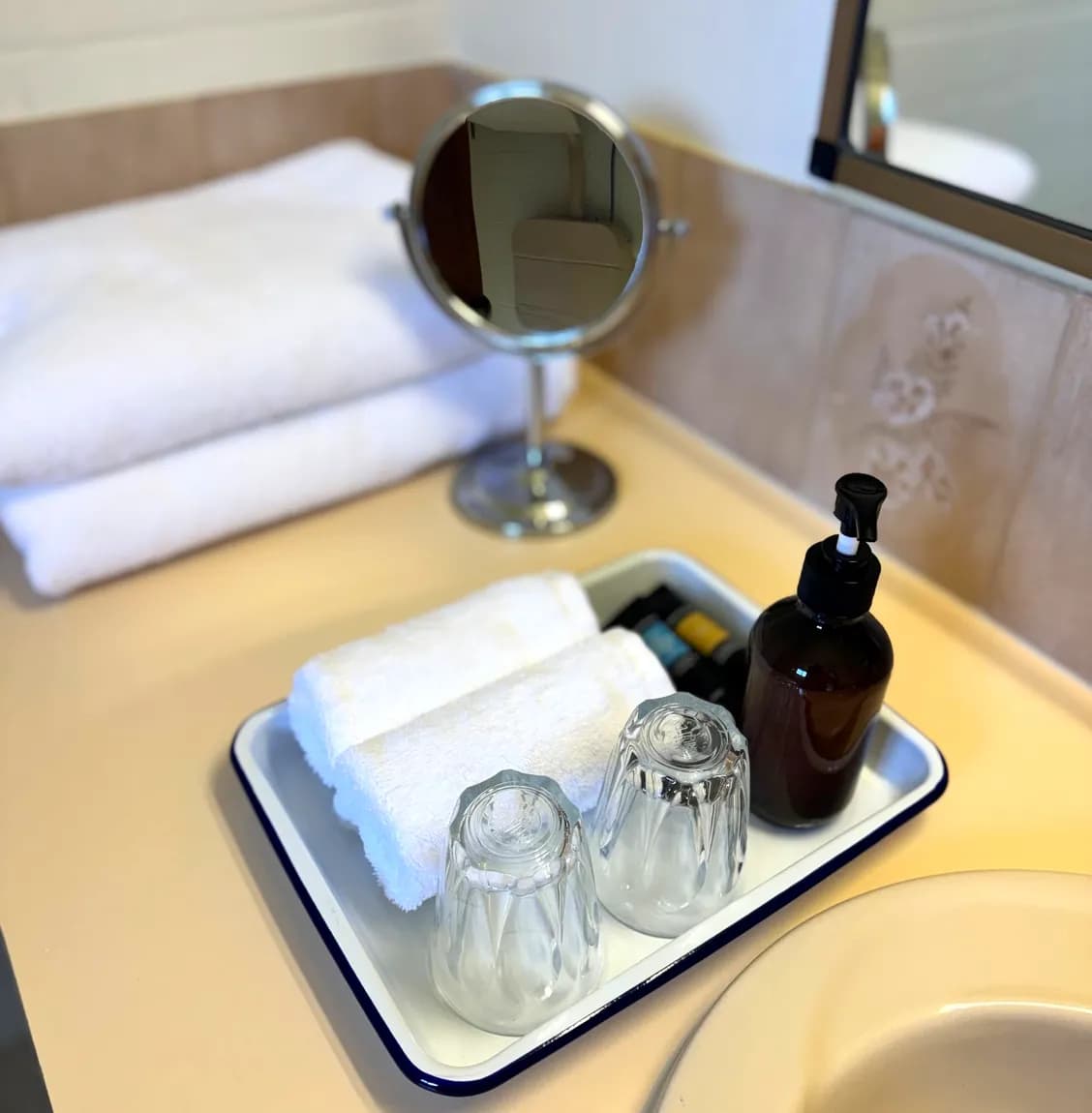 Bathroom tray with folded white towels, two glasses and a bottle of hand wash beside the basin.