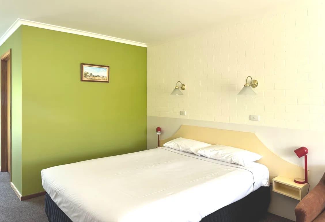 Queen bed with white linen against a green feature wall, a red bedside lamp and a framed print.