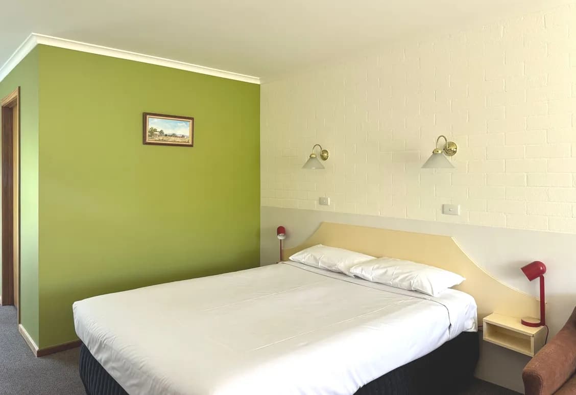 Queen bed with white linen against a green feature wall, a red bedside lamp and a framed print.