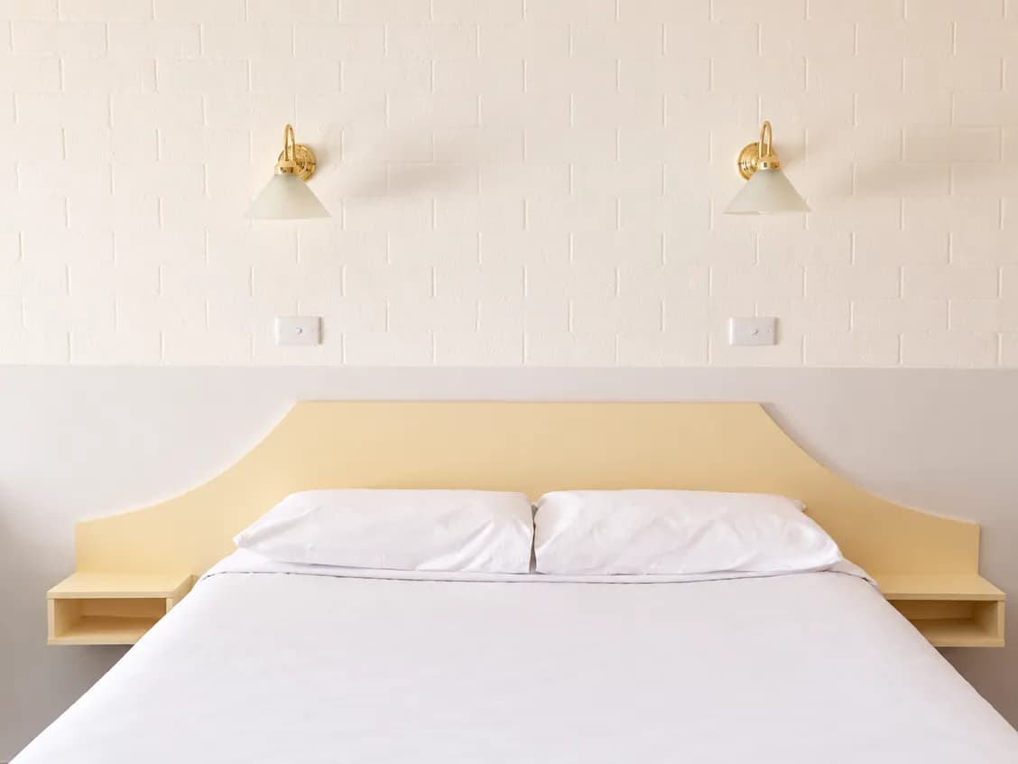 Bed with white linen and a cream bedhead, two brass wall lamps above it on white brick.