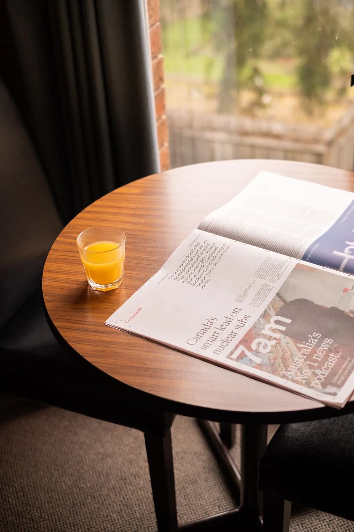 A round wooden table with a newspaper and a glass of orange juice near a window with curtains.