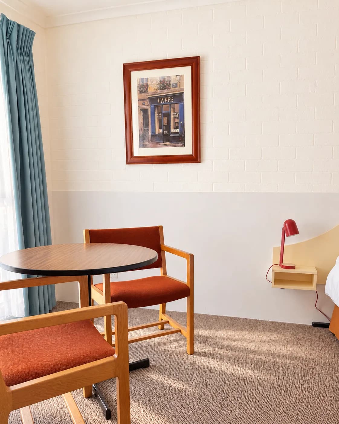 Round table and two orange upholstered chairs by the window, with a framed print and a red lamp on the bedside shelf.