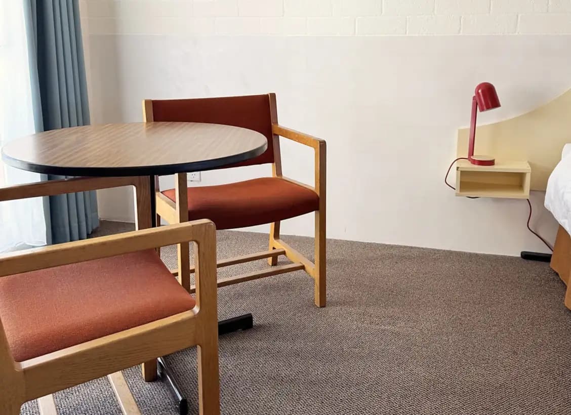 Round table and two upholstered chairs by the window, with a red lamp on the floating bedside shelf.