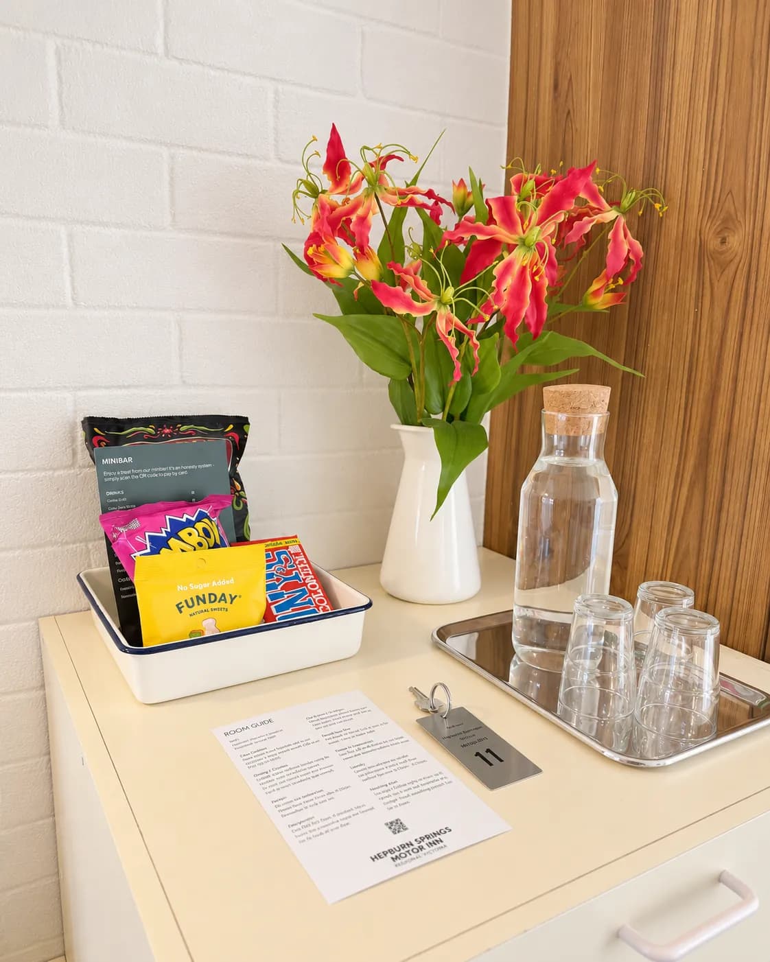 Red lilies in a white jug beside a carafe of water, glasses on a tray, snacks, the room guide and the room key.