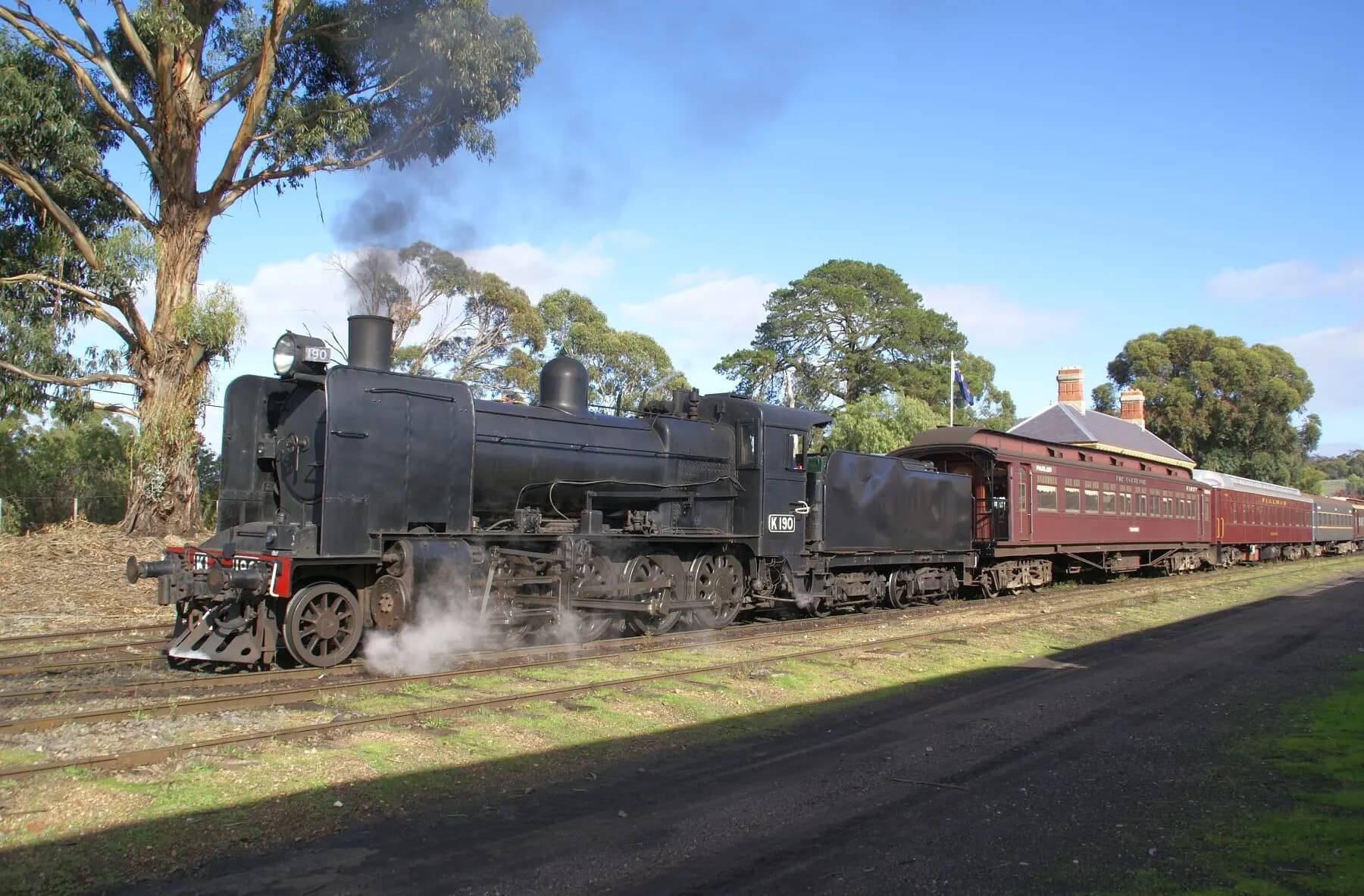 A black steam locomotive, K190, hauling maroon carriages past gum trees on the Victorian Goldfields Railway.