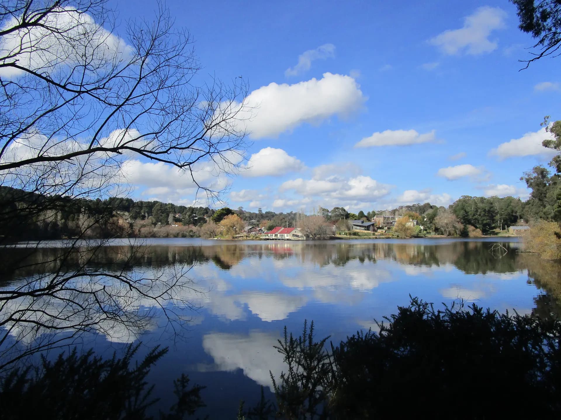 Lake Daylesford on a clear day, the red roof of the Boathouse reflected in still water.